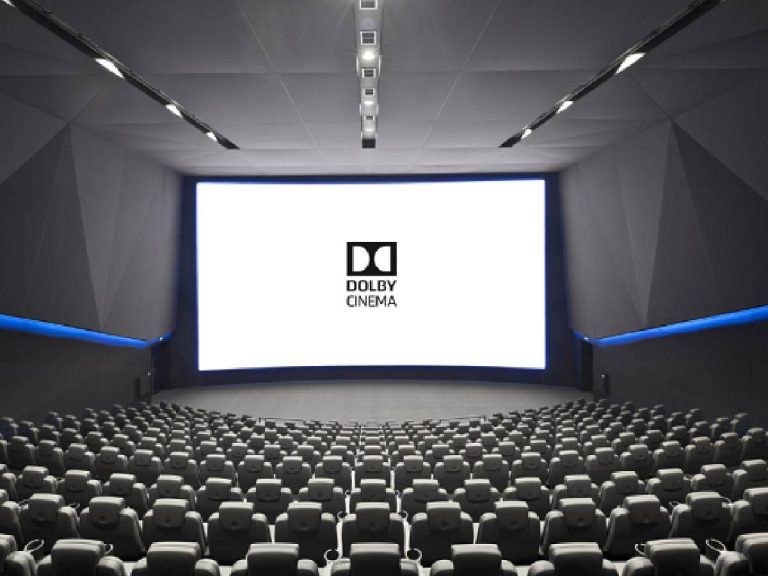 A modern cinema interior featuring a large, bright screen displaying the "Dolby Cinema" logo. The auditorium is designed with a sleek, angular aesthetic and is filled with rows of plush, reclining seats. The walls are adorned with geometric patterns, and there is subtle blue lighting along the edges, enhancing the contemporary atmosphere. The overall setting conveys a high-end movie-watching experience.